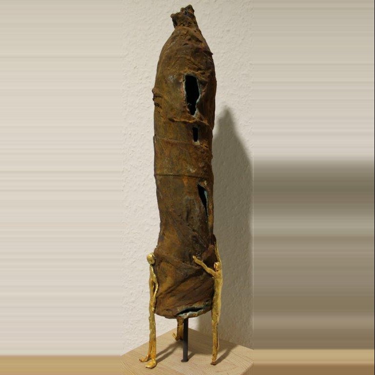 Schlanke, säulenartige Skulptur in Ocker- und Brauntönen mit schmalen Durchbrüchen; am Fuß zwei kleine goldene Figuren, auf einem Holzsockel.